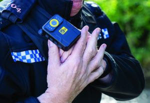 Officer in uniform with blue-white checkered patches holds shoulder-mounted body camera with both hands; device features lens and buttons.