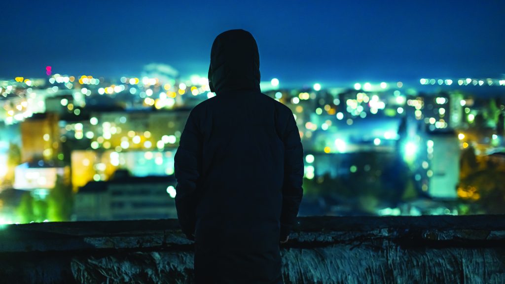 A hooded silhouette stands at a rooftop ledge at night, overlooking a wide cityscape filled with glowing buildings and scattered lights stretching into the distance