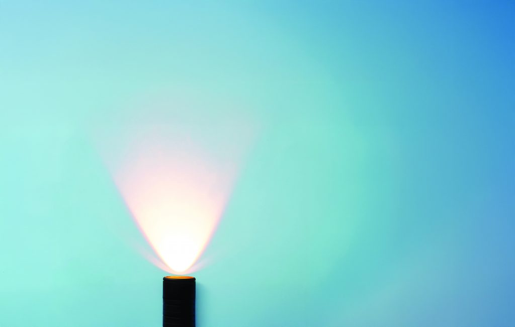 A small flashlight on a surface projects a bright, fan‑shaped beam upward against a smooth blue wall, creating soft gradients and illuminated arcs of light.