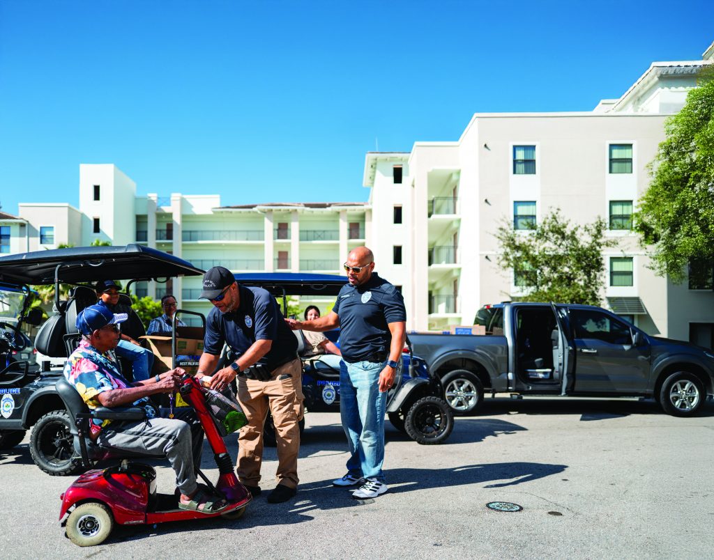 Two law enforcement officers assist a person on a red mobility scooter in a residential area; parked trucks and golf cart visible near multi-story buildings.