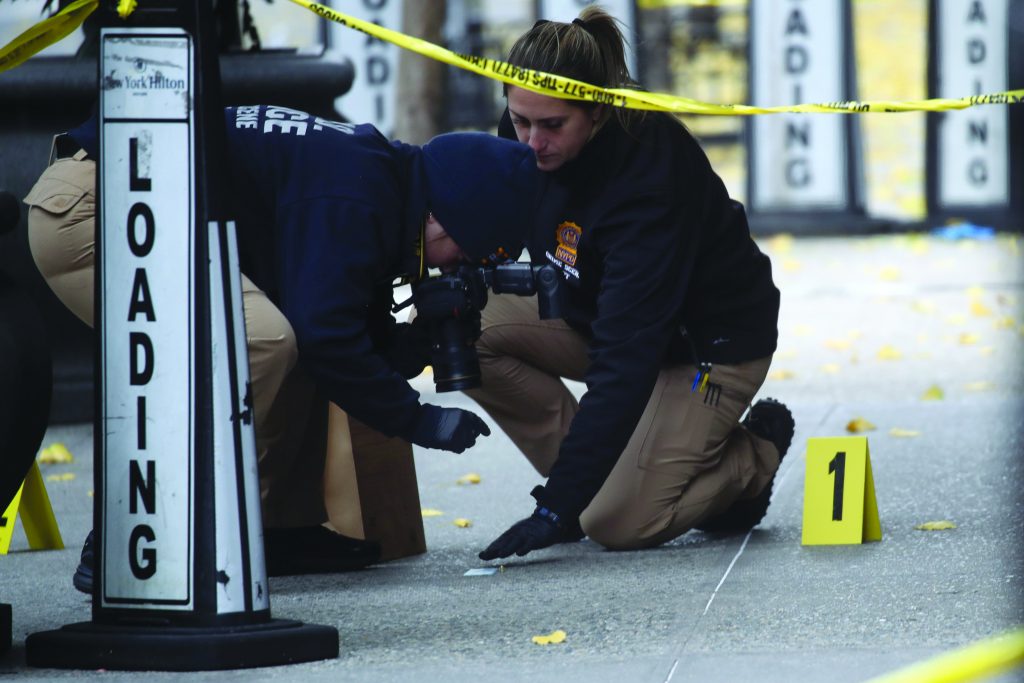 Two officers process crime scene near “LOADING” sign; one kneels by evidence marker “1,” the other holds camera; yellow tape marks perimeter.