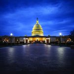 Illuminated U.S. Capitol building at night with glowing dome and columns, set against a dark blue sky, viewed from a wide stone walkway in the foreground
