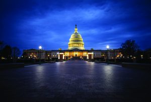 Illuminated U.S. Capitol building at night with glowing dome and columns, set against a dark blue sky, viewed from a wide stone walkway in the foreground