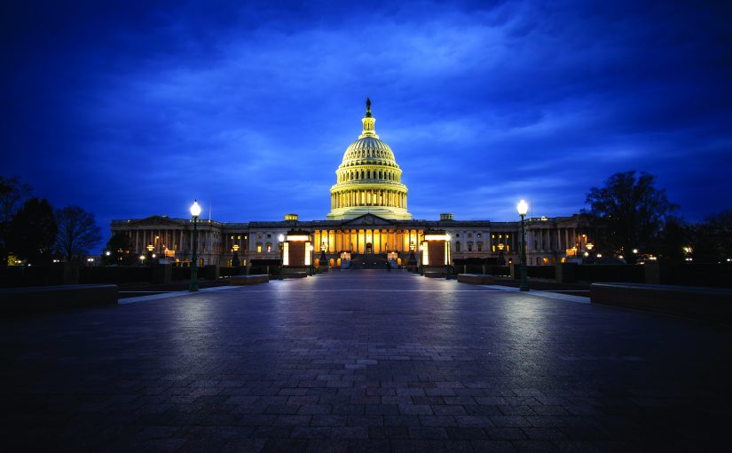 Illuminated U.S. Capitol building at night with glowing dome and columns, set against a dark blue sky, viewed from a wide stone walkway in the foreground