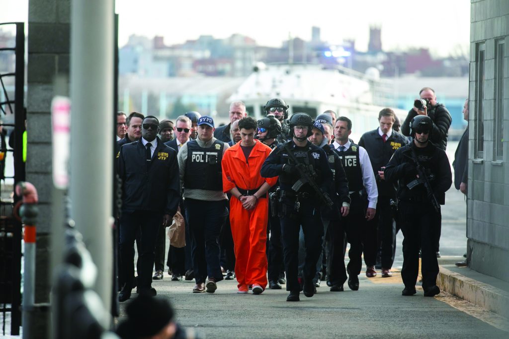 Officers in police and tactical gear escort handcuffed individual in orange jumpsuit; group walks near docked boat, suggesting secure transport.