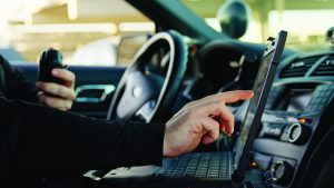 Close-up of a person inside a patrol vehicle using a rugged laptop with one hand while holding a radio, dashboard controls and steering wheel visible