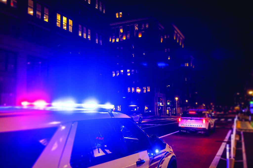 Police SUVs with flashing lights line a city street at night, illuminating tall buildings and traffic signals as they respond to an active scene
