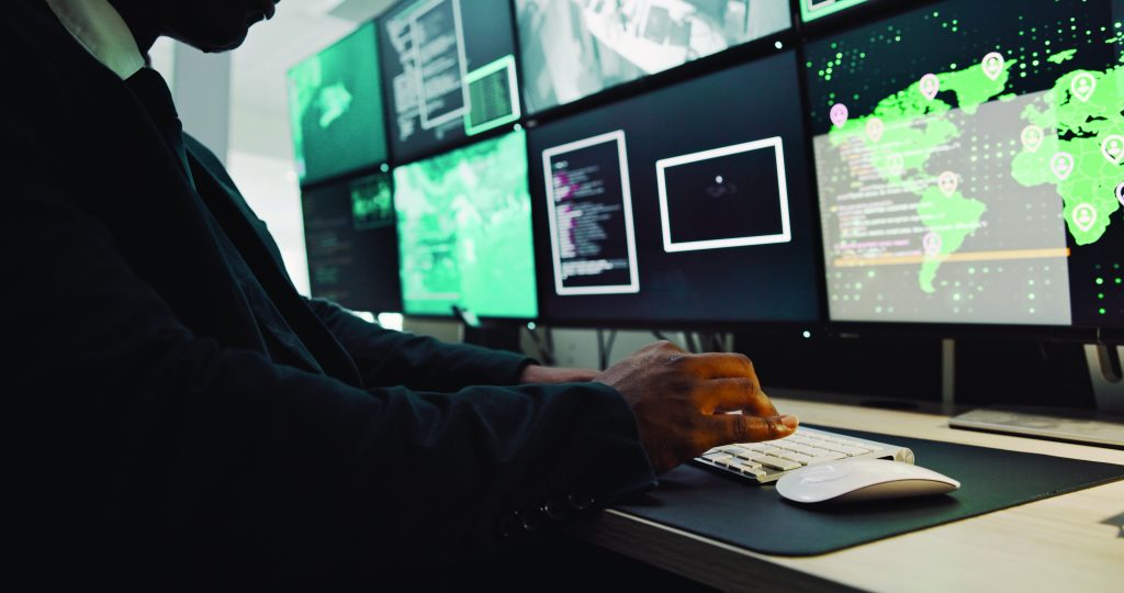 Person in suit at desk with multiple monitors showing maps, data, and surveillance feeds in high-tech control room, highlighting analytical operations