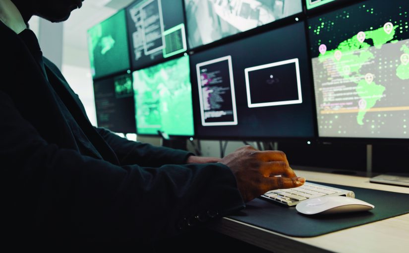 Person in suit at desk with multiple monitors showing maps, data, and surveillance feeds in high-tech control room, highlighting analytical operations
