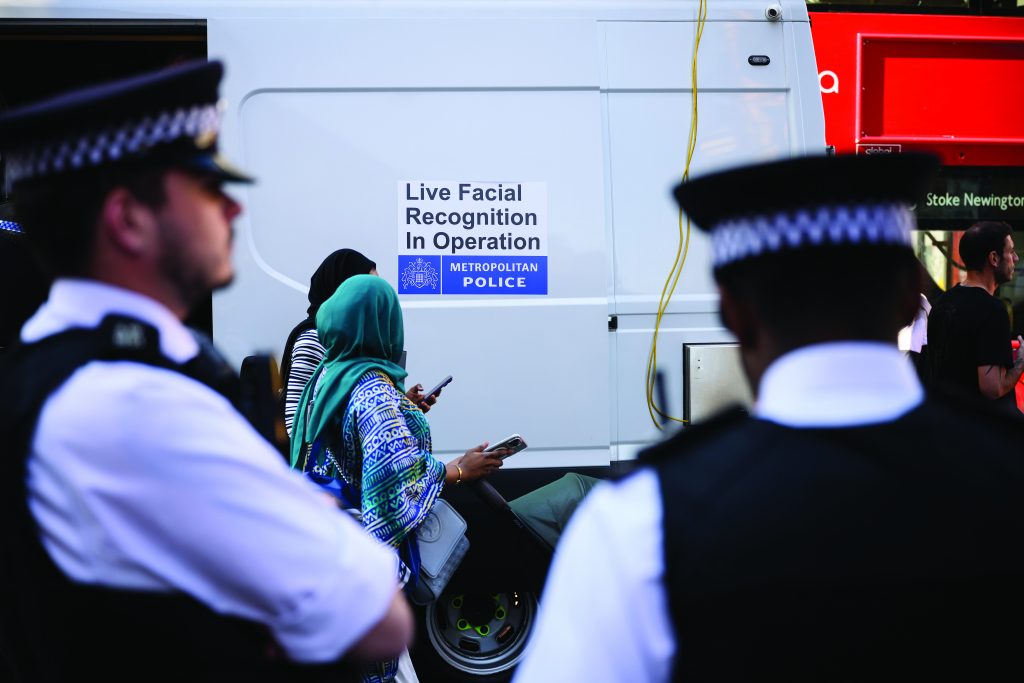 Uniformed police stand near a white mobile unit displaying a sign reading ‘Live Facial Recognition In Operation,’ as members of the public pass along a city street