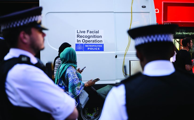 Uniformed police stand near a white mobile unit displaying a sign reading ‘Live Facial Recognition In Operation,’ as members of the public pass along a city street