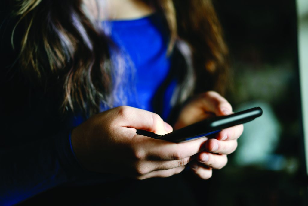 Close-up of hands holding a smartphone, thumbs positioned for typing, with softly blurred indoor background, long hair visible, and blue clothing beneath warm light