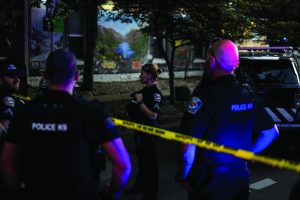 Police officers stand near yellow crime scene tape and a patrol SUV at night, illuminated by blue and red lights, with a mural and trees in the background