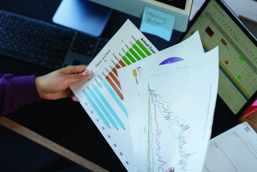 Close-up of a person holding printed charts with colorful bar and line graphs at a desk, next to a keyboard, monitor, and laptop displaying spreadsheets