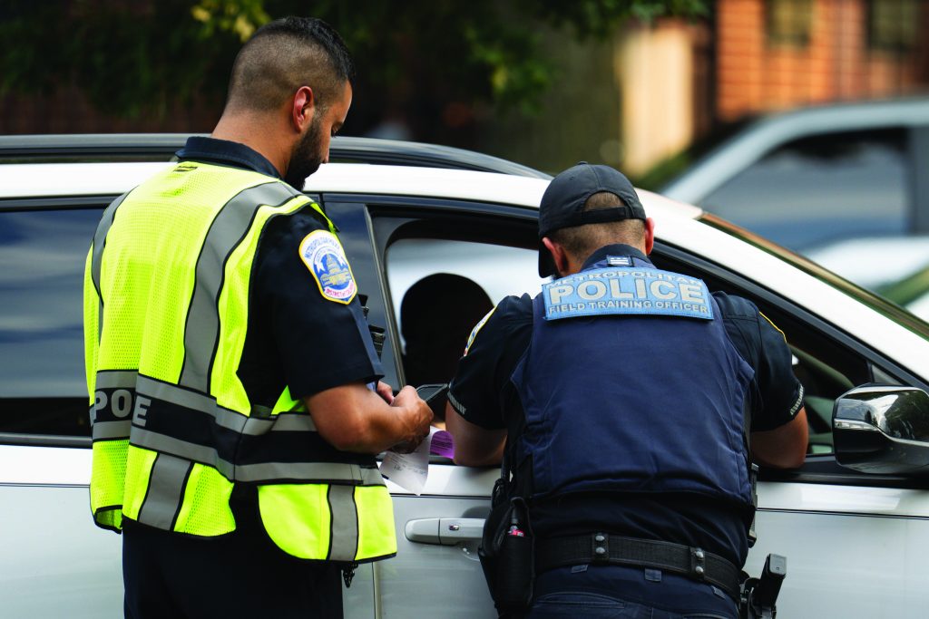 Two officers stand beside a white vehicle during a traffic stop, one wearing a high‑visibility vest holding a ticket while another leans into the driver window