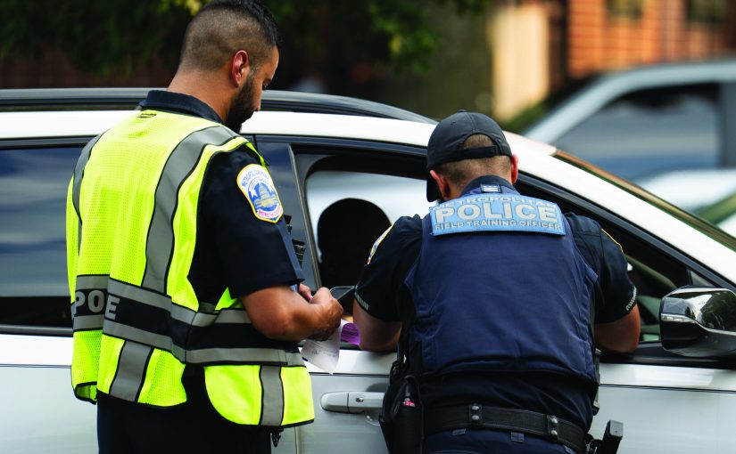 Two officers stand beside a white vehicle during a traffic stop, one wearing a high‑visibility vest holding a ticket while another leans into the driver window