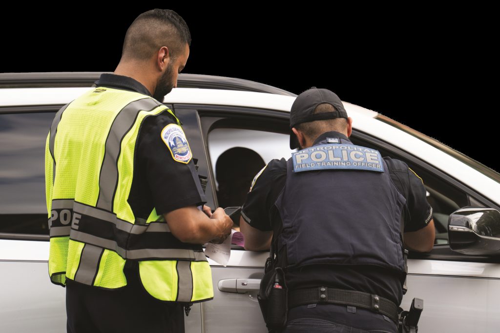 Two police officers conduct a vehicle stop, one in high‑visibility vest writing on a notepad while another leans into a car window, with duty belts, patches, and side mirror visible