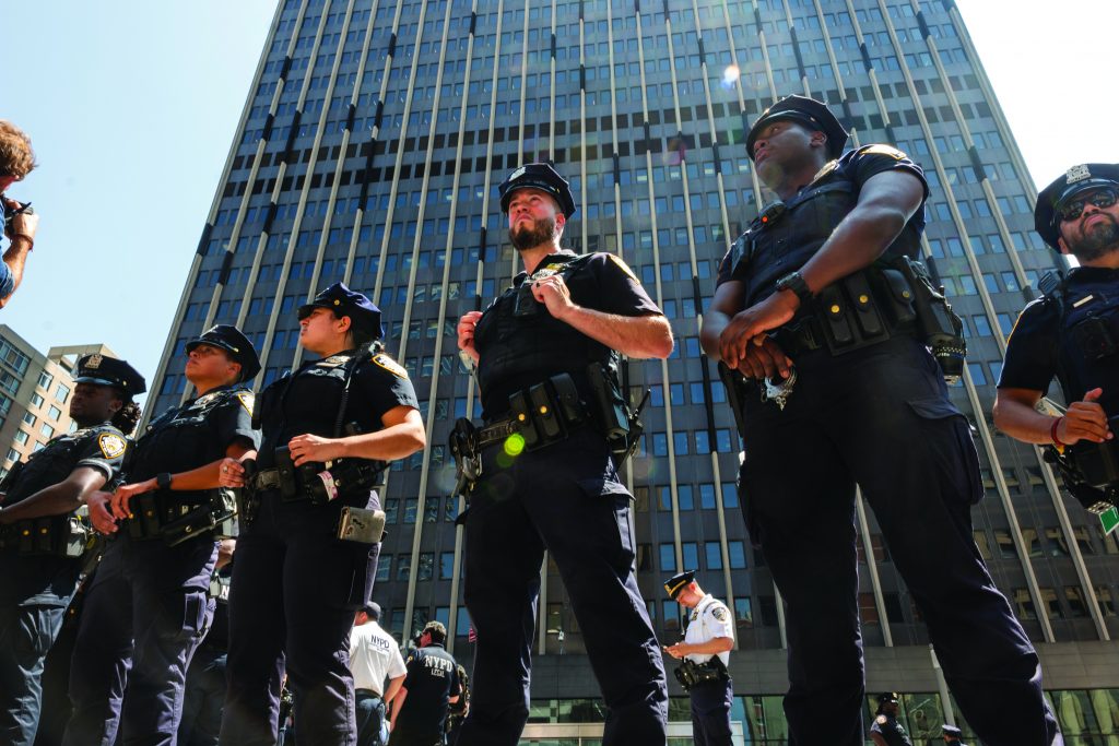 Line of uniformed police officers standing in formation with tactical belts and gear in front of a tall glass office building during daytime.