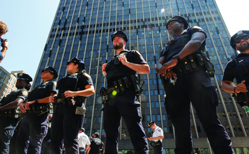 Line of uniformed police officers standing in formation with tactical belts and gear in front of a tall glass office building during daytime.
