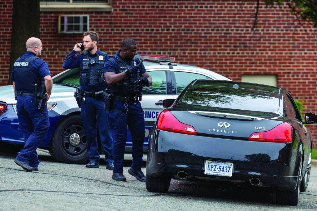 Three officers in body armor and duty belts stand beside a marked Prince George’s County Police cruiser and a black Infiniti coupe, with emergency lights, radios, tactical vests, license plate, and brick apartment building visible