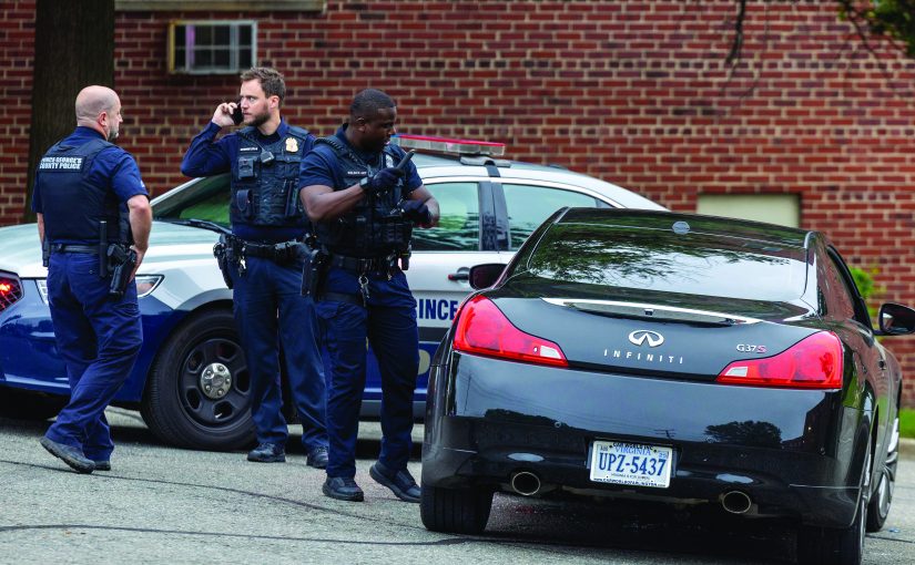 Three officers in body armor and duty belts stand beside a marked Prince George’s County Police cruiser and a black Infiniti coupe, with emergency lights, radios, tactical vests, license plate, and brick apartment building visible