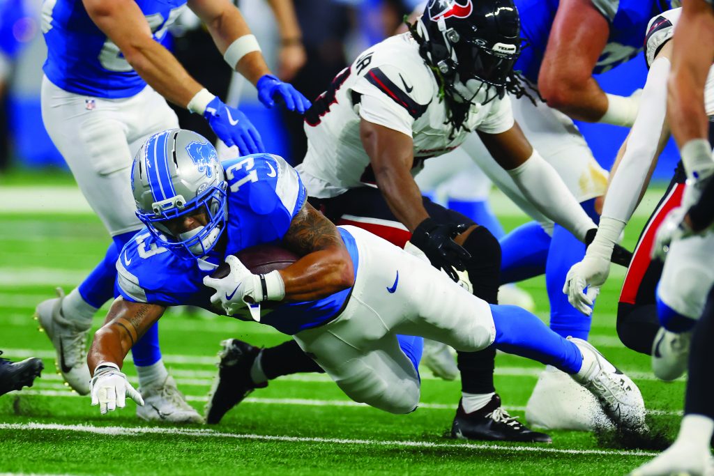 Football player in blue uniform dives forward clutching ball as opponent in white and black uniform tackles from behind on grassy field.