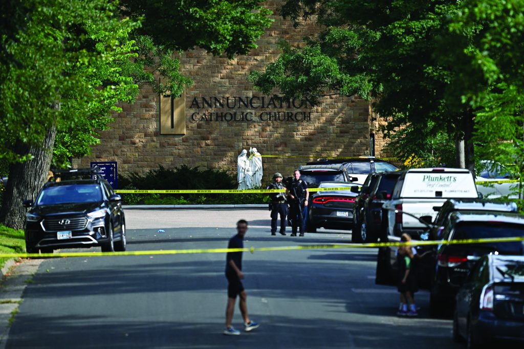 Police vehicles and officers stand behind yellow tape outside a stone church with statues by the entrance as people observe from the street lined with parked cars