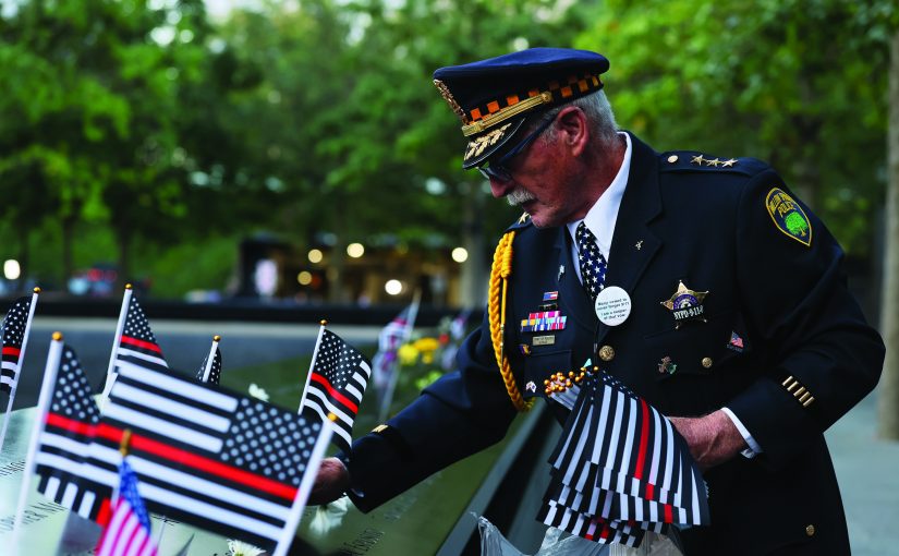 Uniformed police official in decorated dress uniform places black‑and‑white memorial flags and flowers along engraved names at the 9/11 Memorial, with peaked cap, service ribbons, shoulder cord, badge, commemorative pins, reflective pool edge, trees, and evening lights visible