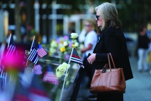 Visitor wearing sunglasses and carrying a tan handbag stands beside an engraved memorial edge adorned with white roses, American flags, and black‑and‑white remembrance flags, while other visitors, trees, flowers, and softly blurred plaza features appear in the background