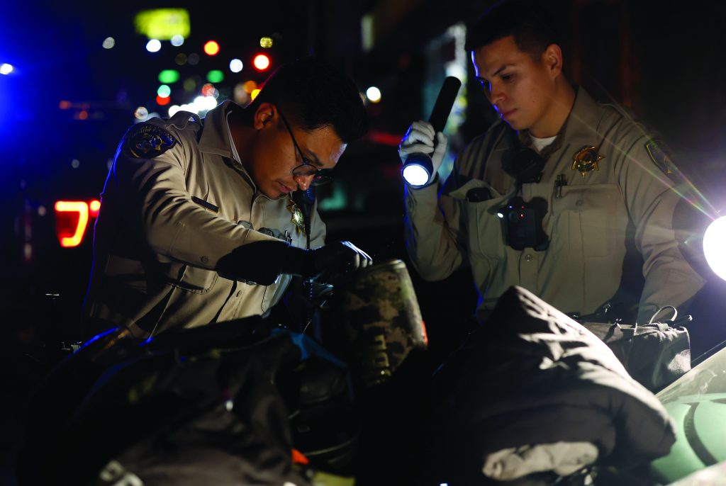 Two uniformed officers inspect items on a vehicle hood at night using flashlights, with tactical gear visible and patrol lights illuminating the scene