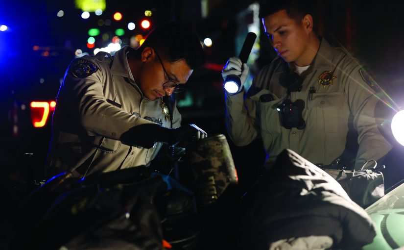 Two uniformed officers inspect items on a vehicle hood at night using flashlights, with tactical gear visible and patrol lights illuminating the scene