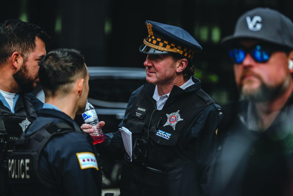 Group of uniformed police officers in tactical vests standing outdoors, one holding a water bottle, badges and patches visible, with vehicles in the background