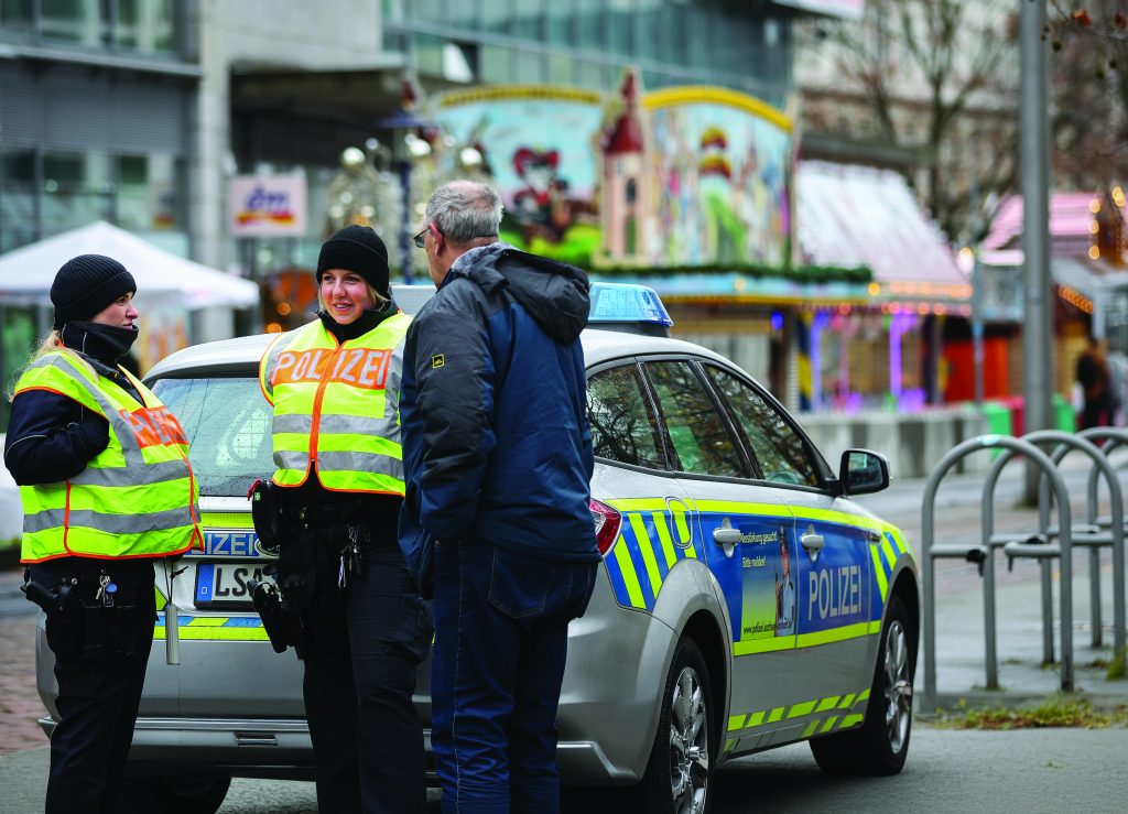 Uniformed police wearing high‑visibility vests stand beside a marked patrol car on a city street, conversing with a civilian near bike racks and storefronts