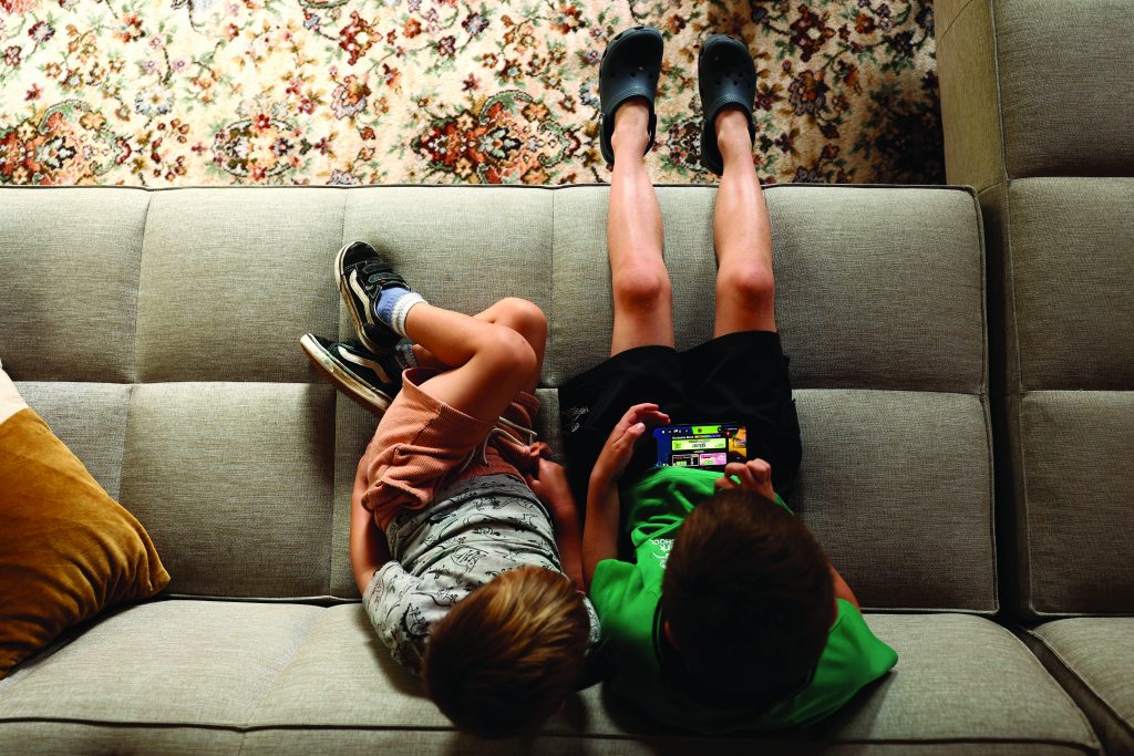 Two children lie on a couch, one playing a game on a handheld device as the other watches, with sneakers, Crocs, and a patterned rug visible above them