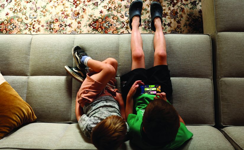 Two children lie on a couch, one playing a game on a handheld device as the other watches, with sneakers, Crocs, and a patterned rug visible above them