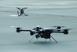 Two drones on a runway, with one grounded and tangled in netting while another stands in the foreground, showing equipment and landing gear on the pavement