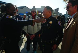 Uniformed police officers greet colleagues and officials on a sunny city street, with badges, shoulder patches, radios, cameras, and bystanders visible among nearby buildings