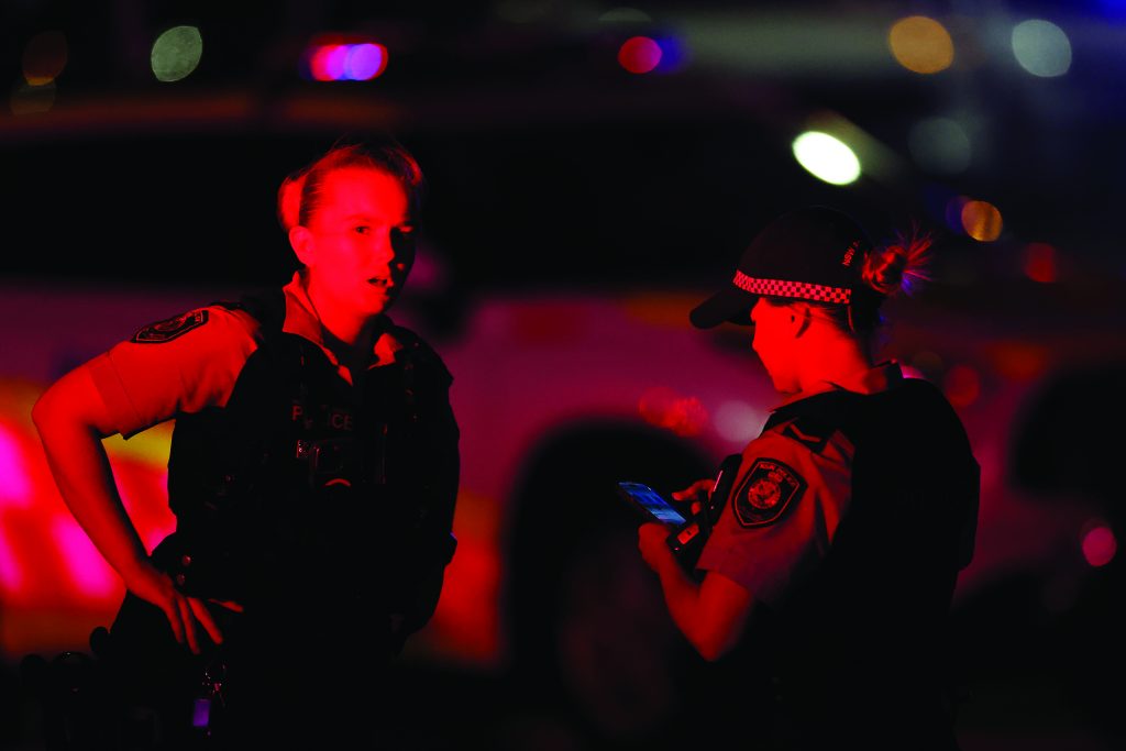 Two uniformed officers stand under red and blue emergency lighting at night, one holding a phone, with blurred patrol vehicles and roadway lights in the background