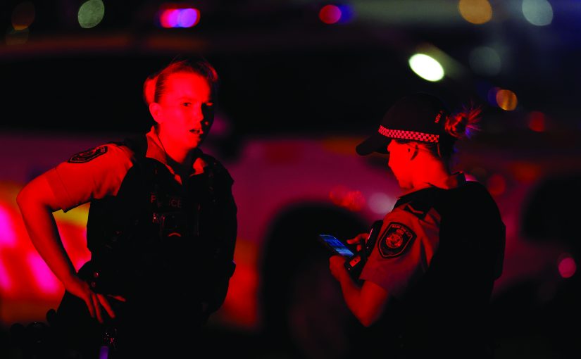 Two uniformed officers stand under red and blue emergency lighting at night, one holding a phone, with blurred patrol vehicles and roadway lights in the background