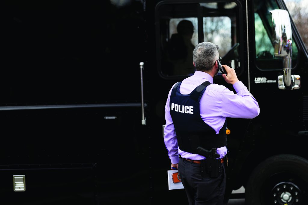 An officer wearing a POLICE‑marked tactical vest speaks into a handheld radio while standing beside a black armored vehicle, holding documents near the rear door