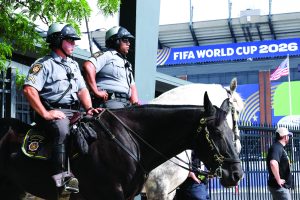 Two mounted Pennsylvania State Police troopers wearing helmets, radios, body cameras, and gray uniforms sit atop black and white horses outside a stadium displaying FIFA World Cup 2026 signage, with fencing, flag, spectators, bridles, reins, and saddle equipment visible