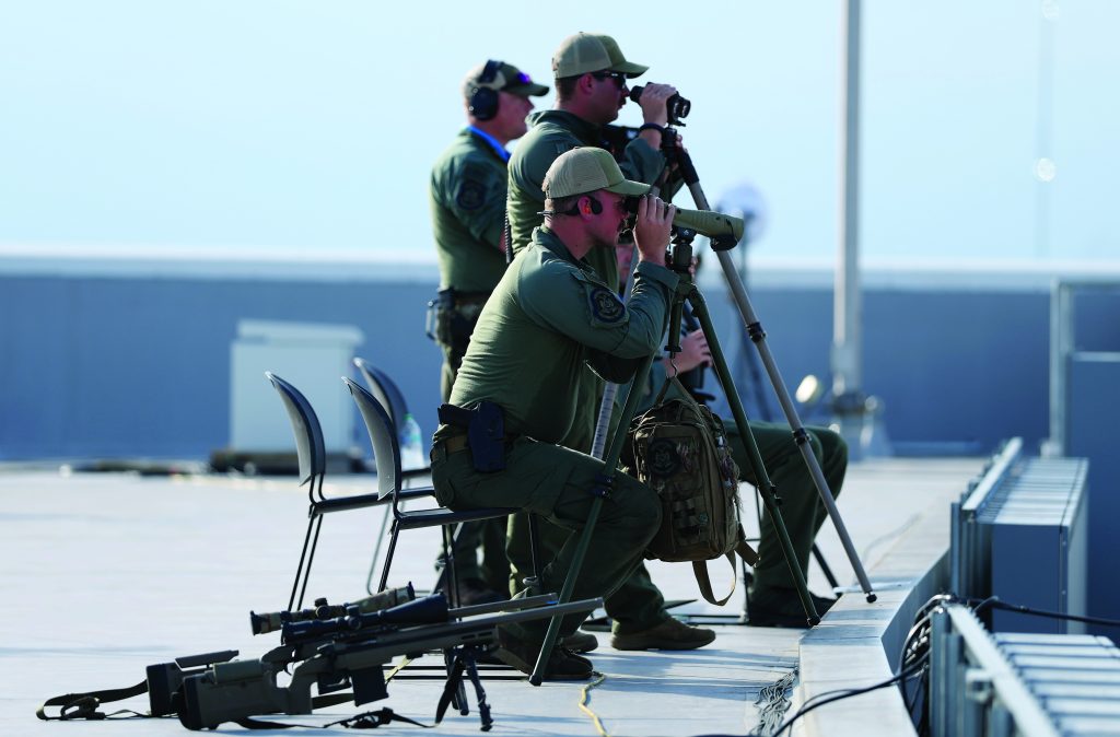 Two mounted Pennsylvania State Police troopers wearing helmets, radios, body cameras, and gray uniforms sit atop black and white horses outside a stadium displaying FIFA World Cup 2026 signage, with fencing, flag, spectators, bridles, reins, and saddle equipment visible.