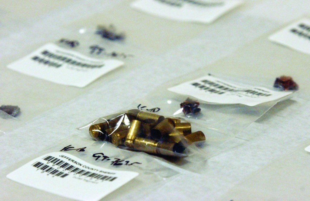 Evidence bags on a table containing spent brass casings and bullet fragments, each labeled with barcodes and handwritten case details for forensic processing