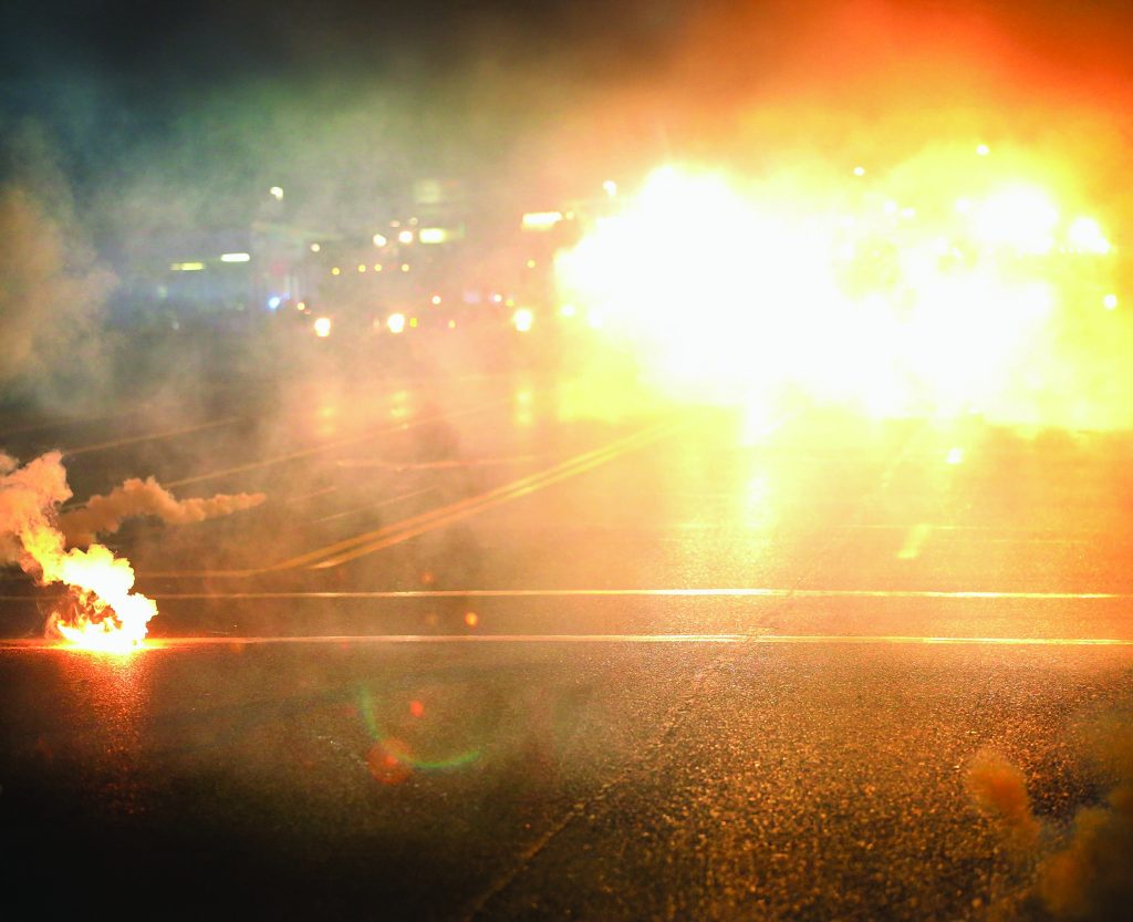Nighttime street scene lit by bright lights and smoke, possibly from flares or tear gas; empty road with lane markings visible through haze.