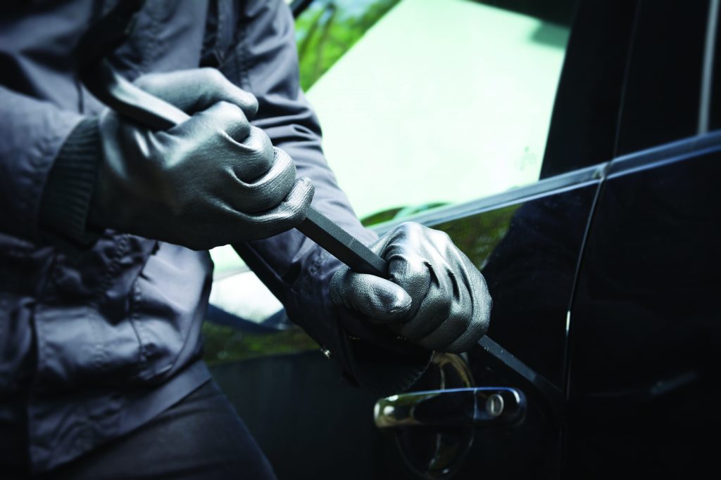 shows a person wearing black gloves and a dark jacket attempting to break into a car using a crowbar. The person's hands are gripping the crowbar, which is being inserted between the car door and the frame near the handle. The car's side mirror and part of the window are visible in the background. The image focuses on the hands, crowbar, and car door, with no faces or other identifying features shown.