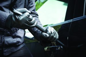 shows a person wearing black gloves and a dark jacket attempting to break into a car using a crowbar. The person's hands are gripping the crowbar, which is being inserted between the car door and the frame near the handle. The car's side mirror and part of the window are visible in the background. The image focuses on the hands, crowbar, and car door, with no faces or other identifying features shown.