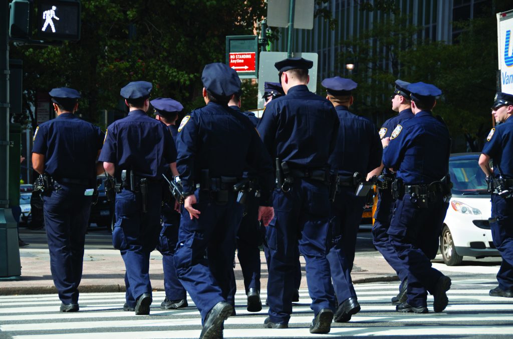 Eight uniformed police officers walk in formation across city crosswalk; pedestrian signal displays “walk” in background; urban setting