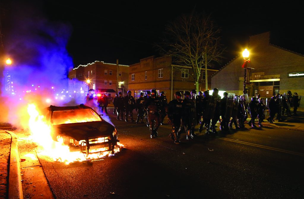 Burning police car on city street at night with flames and smoke; law enforcement in riot gear march past; buildings and streetlights in background.