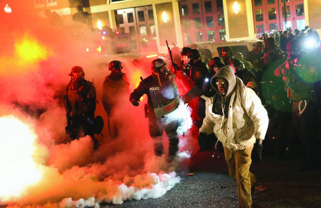Police in riot gear with shields and batons advance through smoke; person in white jacket walks away; tense urban scene with low visibility.