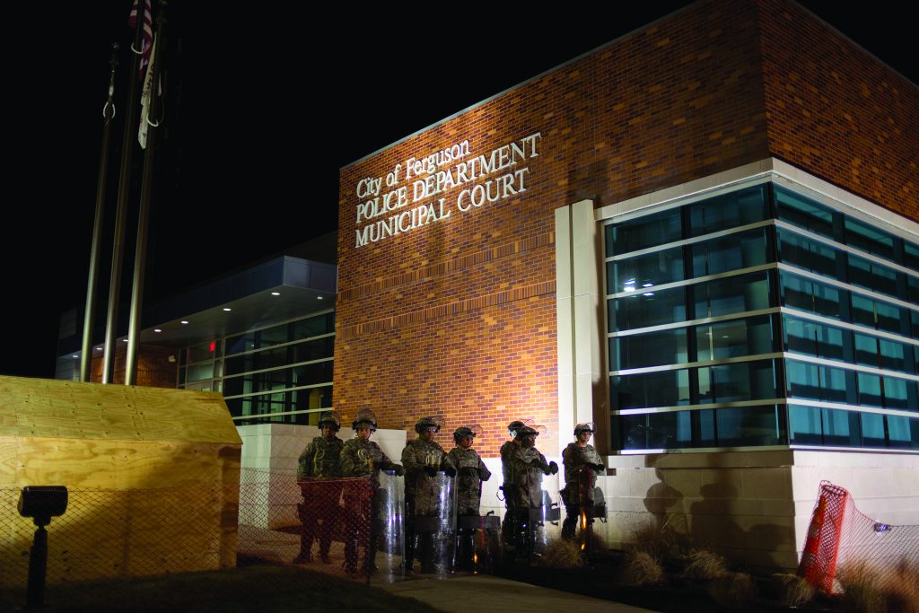 Seven uniformed officers with shields stand at night outside City of Ferguson Police Department and Municipal Court; flags and wooden structure nearby